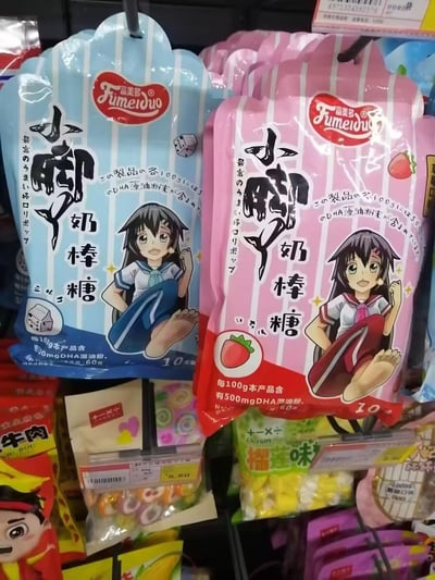 这是一张线下超市货架的实拍图，展示了富美多品牌的「小脚丫奶棒糖」的两种包装：
1. 蓝色包装：带有牛奶盒标识，应为牛奶口味，包装上印有蓝白水手服的动漫女孩，搭配中日双语文字，标注每100g含500mg DHA藻油粉末，净含量60克共10支；
2. 粉色包装：带有草莓标识，为草莓口味，包装上印有红白水手服的同款动漫女孩，同样标注了DHA藻油成分，净含量规格一致。
包装设计以露出脚丫的动漫女孩呼应产品「小脚丫」的命名，风格活泼可爱，面向喜爱零食的消费者，尤其是儿童和喜欢可爱风格的人群。图中还能看到货架上的其他零食，如牛肉零食、榴莲味零食等。