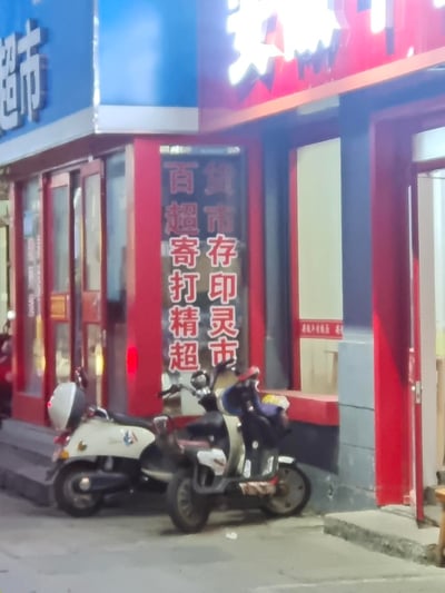 这是一张夜晚拍摄的城市街角街景照片，画面主体是一家红蓝色调的综合服务店铺，店铺玻璃上用红色字体标注了“百货、超市、寄存、打印、精灵、超市”的业务内容，显示出店铺的多元服务属性。店铺门前的人行道上停放着两辆电动自行车，整体场景充满了生活化的烟火气，展现了普通城市夜晚的日常街景。