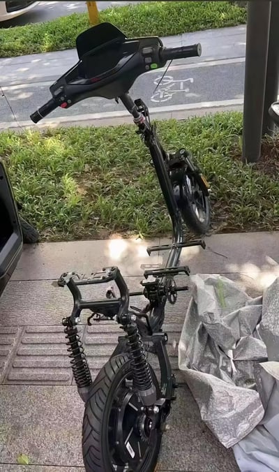 图片展示了一辆黑色电动滑板车倒在人行道上，车身疑似发生拆解或损坏，车架、弹簧、车轮等部件暴露在外。滑板车旁边放着一个灰色的布或塑料袋，背景可见路边的草丛、自行车道标识及停放的车辆，整体场景为日常街头景象，未出现人物或文字信息。