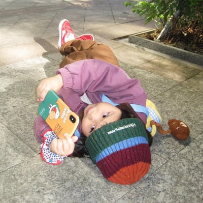 一位穿着彩色毛线帽和休闲服饰的小孩无所谓地躺在地上，双手拿着手机沉迷其中，丝毫不顾周围环境，展现出一种“摆烂”态度。这张图片很适合用来表达心累、摆烂、懒惰、放松自我的状态，也常被网友用在摸鱼和佛系相关的场景。画面没有明显的双关或谐音梗，但整体气质颇具反差萌和沙雕氛围。