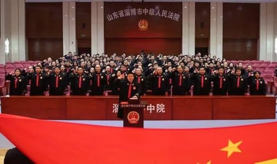 山东省淄博市中级人民法院内举行的法官宣誓仪式现场。画面中，众多身着黑色法官制服、佩戴红色领带的法院工作人员整齐站立，右手举拳进行宣誓。前方设有主席台，背景墙上悬挂着法院名称和徽章，前景可见中国国旗一角。整体场景庄重严肃，展现了司法人员宣誓的正式场合。