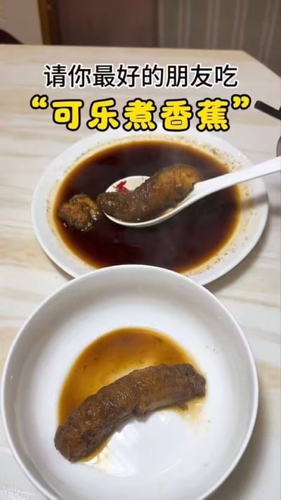 这是一张展示猎奇黑暗料理的实拍图片，画面背景是浅色的桌面，摆放着两个白色餐具：靠后的碗中盛有深褐色的可乐汤汁，汤汁里泡着两段被煮得发黑软烂的香蕉，其中一段被盛放在白色汤勺上；靠前的盘子里也摆着一段可乐煮过的香蕉，旁边残留少量浅褐色汤汁。图片上方配有文字“请你最好的朋友吃‘可乐煮香蕉’”，整体风格搞怪，意在调侃这种口味猎奇的黑暗料理可以用来整蛊好友。