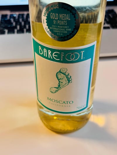 这是一张近距离拍摄的Barefoot（赤足）品牌加州莫斯卡托白葡萄酒的酒瓶实拍图，瓶中装有淡金黄色的酒液。瓶身主标签清晰展示了品牌名“BAREFOOT”、标志性的赤足图案，以及酒款名“MOSCATO CALIFORNIA”和容量“750mL”。瓶身上方的圆形认证标识标注了这款酒在2021年美国蒙特雷国际葡萄酒大赛中斩获金奖，获得91分的评分，还带有“始终如一的品质，公认的价值”的环形文字。背景中露出部分键盘，显示拍摄场景大概率在桌面环境下。