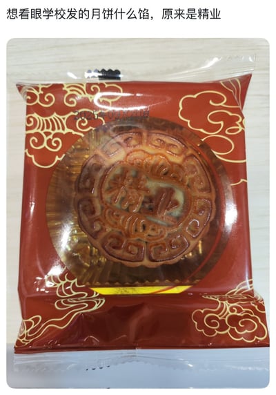 这是一张图文混排的玩梗图，图中展示了一块独立包装的月饼，是学校中秋节发放的福利月饼；包装为红色底色，印有金色祥云纹样，中间设置了透明开窗，可以看到月饼本体，月饼表皮压印有“精业”两个汉字；图片顶部配文“想看眼学校发的月饼什么馅，原来是精业”，玩了文字趣味梗：本意是想要好奇月饼是什么馅料，看到月饼上印的“精业”字样后，就将其作为“馅”的答案，利用文字双关制造了趣味性效果。