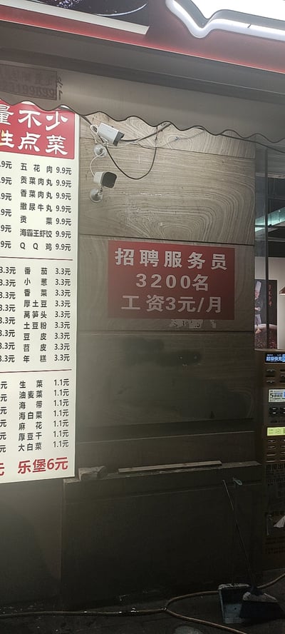 这是一张实拍的餐饮门店内部墙面照片，墙面左侧是该店的菜品价目表，标注了不同品类菜品的亲民售价。墙面右侧贴有一张红色的招聘启事，出现了非常离谱的排版失误，将原本应该是“招聘服务员若干名，工资3200元/月”的信息错写为“招聘服务员3200名，工资3元/月”，内容完全不符合现实常理，产生了强烈的搞笑效果。