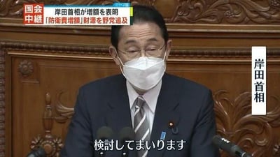 这是一张日本国会会议的直播画面截图，图中人物为岸田文雄，他佩戴白色口罩，身着深色西装搭配条纹领带，坐在带有精致木质雕刻的背景前，面前设有麦克风，正在进行发言。画面左上角有红色的「国会中継」（国会直播）标识，上方的日文标题显示「岸田首相が増額を表明 「防衛費増額」財源を野党追及」，画面右侧有竖排文字标注「岸田首相」，底部的日文字幕为「検討してまいります」，意为“我们将进行探讨”。