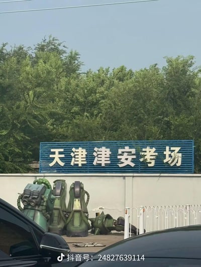 这张图拍摄的是天津津安考场的外部场景，白色围墙上方的蓝色标牌写着“天津津安考场”，围墙下摆放着几个《机动战士高达》系列里的扎古机甲模型，其中一个模型倒地，其余的立在一旁。这个场景充满诙谐的反差感，很容易让人联想到考驾照挂科的考生，仿佛倒地的扎古就是没通过考试的“考生”，用来调侃驾照考试的难度与挂科的心情。