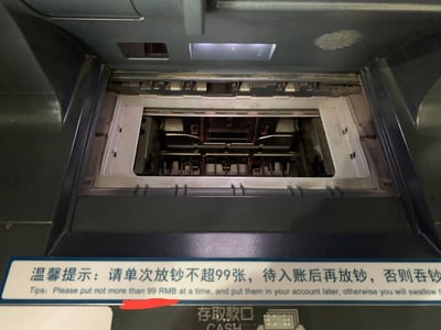 这张图片实拍了银行ATM机的存取款口区域，内部可见用于识别钞票的机械结构，存取款口下方贴有蓝底白字的双语温馨提示牌，中文提示用户单次放钞不得超过99张，需等待入账完成后再继续放钞，违规操作可能导致吞钞；英文翻译对应提示相关注意事项，英文内容末尾有截断。