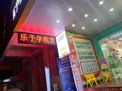 这是一张夜间拍摄的线下少儿教育培训机构门店照片，门店名为「乐予学教育」，定位为青少年素质中心，提供少儿美术、少儿书法等课程。门店入口处设有红色电子发光招牌，门口摆放多座宣传展架，分别宣传英语家长培训课程、购课优惠活动；店内墙面展示了学员作品照片，搭配黄色小椅子，天花板装有多盏嵌入式射灯，整体呈现出面向少儿的课外培训场所的正式宣传氛围。