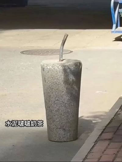 这张图拍摄于户外水泥人行道上，主体是一个圆柱形灰色石质路障，路障顶部插有一根弯曲的金属杆，整体外形和插了吸管的奶茶杯高度相似，图片左下角配文“水泥啵啵奶茶”，用幽默的方式将普通路障恶搞成了新型奶茶，外形巧合带来的反差感十足，充满趣味。