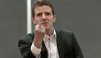 图片中是一位穿着黑色西装和白色衬衫的男性，他正对着镜头做出竖中指的手势。他的表情严肃，眼神直视前方，衬衫上别着一个麦克风，背景是灰色的墙壁。这个表情包通常用来表达愤怒、不屑或反抗的情绪。