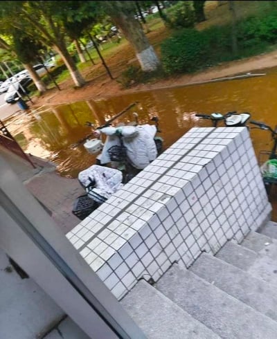 暴雨过后，一处积水严重的路段，一名骑摩托车的人正艰难地试图通过浑浊的黄色积水。画面从高处台阶向下拍摄，可见积水已没过摩托车轮胎，骑车人身后还载有物品。周围有树木和停放的车辆，显示这是一个有绿化的城市道路场景。