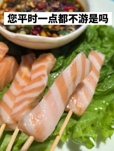 整张图的主体是数串被穿在竹签上的三文鱼腩，鱼肉脂肪纹理丰厚明显，被摆放在新鲜的生菜叶上，图片背景处还有一碗混合了辣椒、葱花、蒜末等配料的蘸料汁。图片上方配文“您平时一点都不游是吗”，是玩调侃的梗，打趣这块三文鱼的脂肪含量过高，仿佛对应的鱼生前完全不游动锻炼才积攒了这么多脂肪，趣味十足。