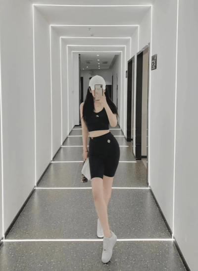 一位穿着黑色运动套装和白色帽子的女性在具有未来感的白色走廊中自拍。走廊采用了LED线条灯设计，创造出透视延伸效果，地面为灰色水磨石材质。女性手持手机遮挡面部，姿态自然，整体风格展现出运动时尚感。