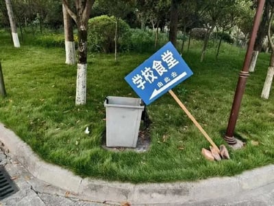 一张校园场景照片：绿色草坪上，一块蓝色的“学校食堂 由此左”指示牌歪斜地倚靠在灰色垃圾桶上，牌面箭头指向左侧。周围有树木、灌木丛和人行道，指示牌的不当放置形成幽默反差，仿佛在“指引”前往垃圾桶而非食堂。