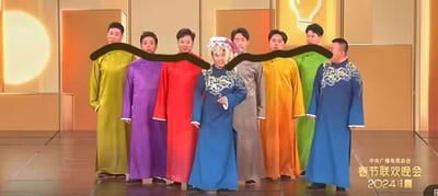 舞台上站着8位表演者，身着绿、紫、红、蓝、橙、灰等多种颜色的传统长衫服饰，背景为暖金色灯光效果。中间一人穿着蓝色绣花纹样服装并佩戴白色头饰，整体呈现出喜庆正式的氛围。画面底部右侧有“中央广播电视总台 春节联欢晚会 2024”水印，人物面部被一条黑色曲线覆盖。