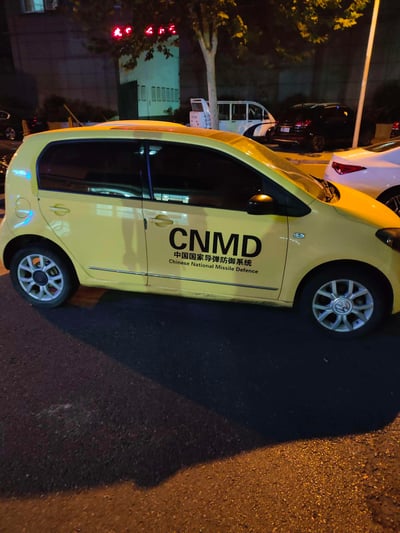 夜晚街道上停着一辆黄色的小型轿车，车身侧面贴有黑色的"CNMD 中国国家导弹防御系统 Chinese National Missile Defence"标识。周围有其他车辆停放，背景有建筑物和树木，营造出一种反差萌的搞笑效果。