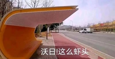 这是一张户外原始拍摄的横版静态照片，画面展示了路边一个造型奇特的黄色公交站台，从拍摄角度看，这个站台的弯曲结构和外形高度酷似虾的头部。图片底部配有文字“沃日!这么虾头”，其中运用了谐音梗，“虾头”谐音“下头”，表达了拍摄者对该公交站台设计的吐槽情绪，认为这个设计十分扫兴、不合常理。