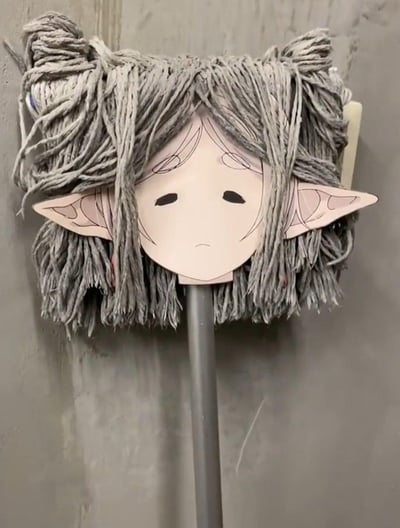图片中展示了一个创意DIY作品：一把灰色的拖把，其纤维部分被巧妙地塑造成凌乱的“头发”造型，拖把头部正中央粘贴着一张纸质卡通脸。这张脸有着尖尖的精灵耳朵、苍白的肤色，以及一双下垂的黑色眼睛和微微下撇的嘴角，整体呈现出明显的悲伤表情。背景是朴素的灰色水泥墙面，使得拖把与卡通脸的组合成为视觉焦点。这种将日常清洁工具与可爱又带点忧郁的卡通形象结合的方式，既幽默又带有一丝反差萌，营造出独特的趣味效果。