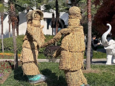 这是一张户外园林场景的实拍照片，画面核心是两个由稻草手工编织而成的拟人艺术装置：左侧造型戴着类似草帽的稻草装饰，二者手臂相牵，姿态仿佛在携手共舞，装置的结构模仿了分层服饰的样式，充满创意。背景中可以看到棕榈类树木、茂密的绿植灌木丛，远处还有白色大象雕塑和行人，整体场景将传统稻草工艺与现代创意结合，营造出温馨有趣的田园氛围。
