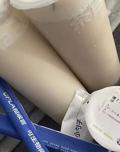 这是一张第一视角拍摄的实景照片，画面主体是拍摄者穿着浅色丝袜的双腿，腿前摆放着茶百道品牌的奶茶相关配件：蓝色包装的CPLA可生物降解粗吸管、印有“小勺子”字样的餐具包装袋，还有贴有订单标签的奶茶杯一角，标签上可见“椰子1份”、茶百道品牌slogan“好茶为底，制造新鲜”等文字。这张图利用物品摆放角度制造了视觉错位效果：长条形的蓝色吸管包装刚好放在腿部前方位置，很容易让观看者产生误解，搭配原作者的标题“想什么呢”，形成了调侃观看者想歪的趣味梗。