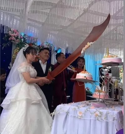 婚礼现场，新娘身着白色婚纱、头戴皇冠，新郎穿着黑色西装，两人共同手持一把造型夸张的巨型刀具，正准备切割桌上的多层婚礼蛋糕。背景装饰有白色流苏和粉色花卉，营造出浪漫喜庆的氛围。画面因使用超大尺寸的刀具切蛋糕而显得十分有趣和独特，新人脸上洋溢着幸福的笑容。