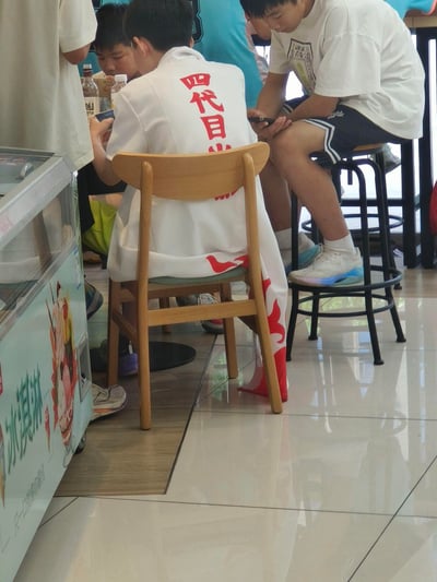 在一家室内零食/饮品小店中，一名男生背对着镜头坐在木椅上，身着印有红色“四代目火影”字样的白色披风，正低头专注操作手机；身旁还有几名年轻男生，或坐或站，同样都在使用手机。画面左侧可见一台标注“冰淇淋 只一口就将你融化”的冷藏柜，整体呈现出年轻人聚集在一起休闲放松的日常场景。