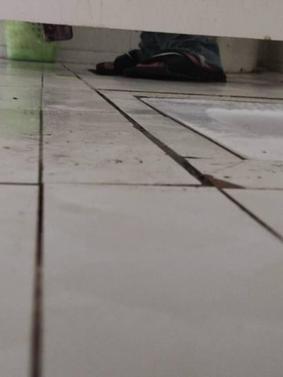 这张图片以低视角拍摄，画面主体是带有深色填缝剂的浅色瓷砖地面，瓷砖表面分布着少量污渍，右上角可见一处类似蹲便器的设施区域；画面上方的柜体下方，露出了穿着深色夹脚拖鞋与蓝色牛仔裤的人的脚部，人物的腿部和身体被柜体遮挡，仅能看到脚部部分。整体场景偏向生活化，推测是在卫生间或厨卫类的室内空间，没有双关、谐音梗或暗示类内容。