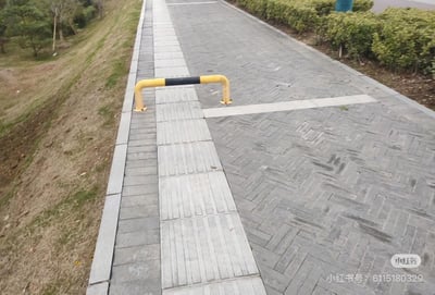 这是一张户外实拍图，画面左侧是长有稀疏草木的斜坡，中间为人行步道，步道左侧铺设有供视障人士使用的长条状盲道，盲道的通行路径正上方被一根黄黑配色的U型阻车栏杆横向完全阻拦，彻底切断了盲道的通行路线，步道右侧是成片的绿色低矮灌木丛，右下角带有小红书平台的水印及发布者ID，该设计完全没有考虑视障人群的出行需求，存在极大的安全隐患。