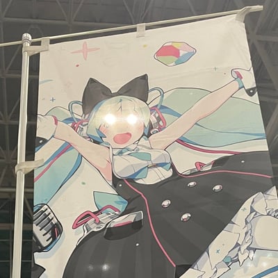 室内展会现场悬挂的动漫风格横幅，画面主体为蓝发猫耳少女，身着白色上衣与黑色短裙，双臂张开呈欢呼姿态，嘴巴大张露出兴奋表情。角色头部佩戴耳机，背景有粉色星星和彩色菱形图案点缀，整体色调明快。横幅因现场灯光照射，在角色面部形成明显反光光斑，展现出展会现场的真实拍摄效果。