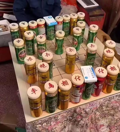 一张创意十足的照片，展示了一副用啤酒瓶代替棋子的中国象棋。绿色和金色的啤酒瓶（主要是青岛啤酒）被整齐地摆放在象棋棋盘上，每个瓶盖都用红色马克笔写上了象棋棋子的名称（兵、炮、相、仕、马、车、帅等）。棋盘上还散落着几盒药品，增添了生活气息。背景中可以看到一位穿着蓝色外套的人，整个场景呈现出一种轻松幽默的生活创意。这种将日常物品巧妙结合传统游戏的方式，展现了普通人的生活智慧和娱乐精神。