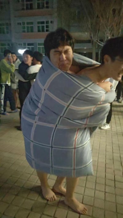 夜晚户外场景中，两名男子被一条蓝色格子毯子紧紧裹在一起，表情夸张滑稽，似乎在进行搞笑互动。背景中可见多名围观者，有人在拍照或录像，整体氛围轻松欢乐，疑似校园或社区内的朋友间趣味活动。