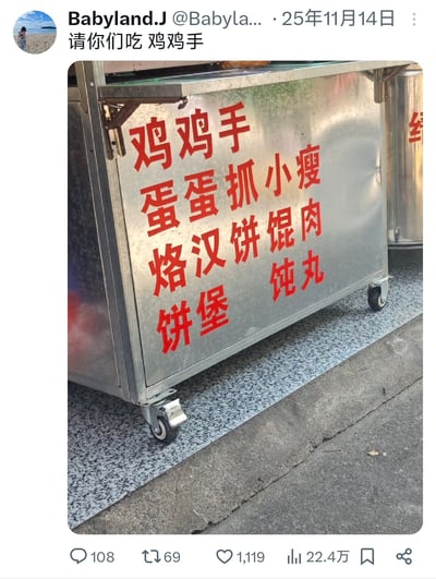一张街头小吃车的照片，车身用红色大字写着各种食品名称："鸡鸡手 蛋蛋抓小瘦 烙汉饼馄肉 饼堡 饨丸"。最引人注目的是开头的"鸡鸡手"和"蛋蛋抓"，这两个词在中文里有明显的双关含义，原本可能想写"鸡爪"和"蛋抓"，却因重复用字意外形成了搞笑的生殖器谐音梗，让人忍俊不禁。小吃车为不锈钢材质，带有轮子，停放在灰色地面上，整体看起来是真实的街头美食摊位。