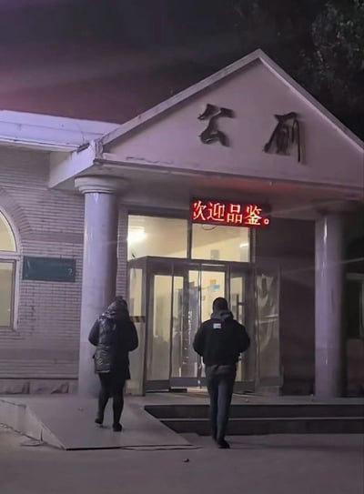 这是一张夜晚拍摄的户外实拍照片，画面主体是一处公厕的入口建筑，建筑门头的三角形墙面上有两个褪色的“公厕”大字，门口上方的红色电子显示屏出现故障，只显示出“次迎品鉴。”五个字，应该是原本的欢迎类标语第一个字显示不全导致的错误。有两名路人背对着镜头，正朝着公厕入口行走，整体是真实生活场景，因错字产生了反差搞笑效果。