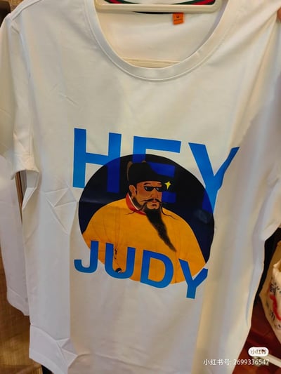 一件白色T恤，正面印有蓝色的"HEY JUDY"文字和一个戴着墨镜的历史人物肖像。历史人物身穿黄色传统服饰，头戴黑色帽子，留有胡须，表情严肃中带着一丝酷感。文字与人物肖像结合，形成了一种古今混搭的幽默效果。