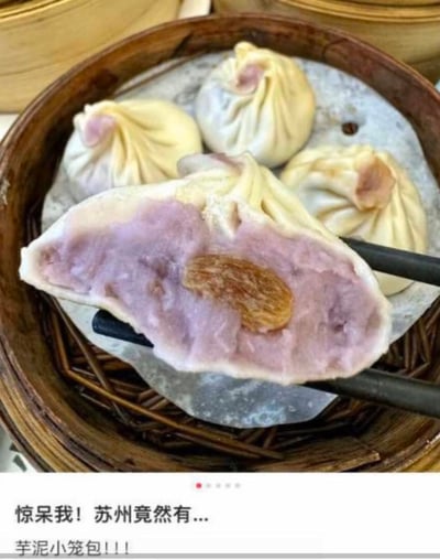 这张图展示了苏州的创新美食——芋泥小笼包：竹制蒸笼里摆放着几个造型和传统小笼包一致的包子，其中一个被筷子夹开，露出粉紫色的绵密芋泥内馅，还嵌有一颗葡萄干，彻底打破了大众对小笼包都是咸口肉馅的固有认知。图片下方配文“惊呆我！苏州竟然有...芋泥小笼包！！！”，传递出对这种新奇甜口小笼包的惊讶与新奇感。