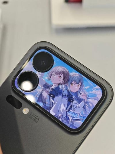 特写展示了一部带有Leica标志的智能手机摄像头模组，镜头保护盖区域印有一幅二次元插画：两名身着校服的少女并肩而立，左侧少女为紫色短发，右眼微眯抬手微笑；右侧少女为蓝色长发，金色眼眸，手指轻触唇边，背景是蓝天白云。手机整体为深灰色，摄像头模组边缘有光泽反射，整体风格清新可爱，融合了科技产品与二次元文化元素。