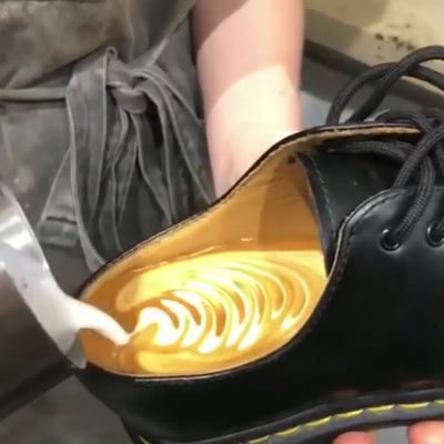图片中展示了一只黑色皮鞋被当作容器，里面盛着金黄色的液体，有人正用奶泡壶在鞋中拉出类似拿铁咖啡的漩涡花纹。皮鞋的鞋带松散地系着，旁边能看到穿着灰色围裙的人的手臂。整个画面充满反差感，将日常用品用于非传统用途，营造出荒诞又有趣的视觉效果。