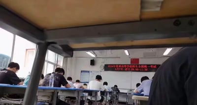 这张图片采用考场座位下方仰拍的特殊视角拍摄，主体为高考考场内部场景：多排课桌前坐满了考生，考生们都低头面向桌面处于答题状态；教室前方墙面悬挂的电子显示屏上标注有“2026年普通高等学校招生全国统一考试 6月7日 语文09:00-11:30”的红色文字，教室内还可见照明灯、挂钟、公告栏、大面积窗户等常规校园教室设施，画面上方露出了前排课桌的底部金属与木质结构，画面右侧有拍摄者附近的衣物局部入镜。
