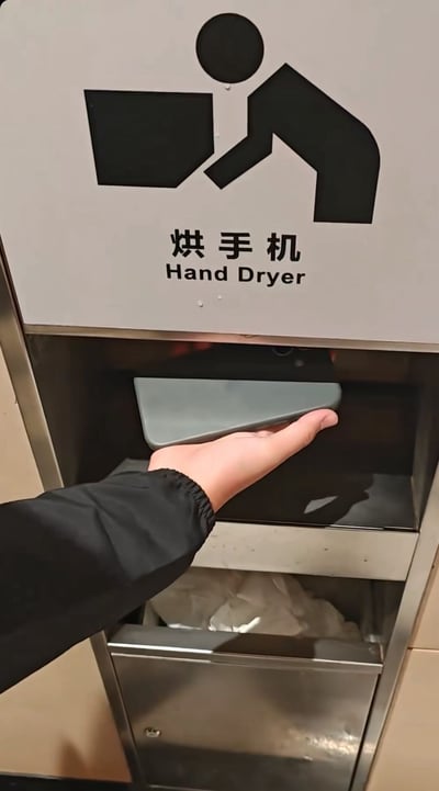 图片展示了公共卫生间的烘手机设备，上方白色指示牌印有黑色人形烘干手部的图标，下方标注中文“烘手机”及英文“Hand Dryer”。一只穿着黑色外套的手正伸向烘手机的银色出风口，似乎准备使用。图中核心笑点在于中文“烘手机”的字面歧义：字面意为“烘干手机”，实际功能是“烘干手部”，这种文字与实际用途的反差形成幽默效果。