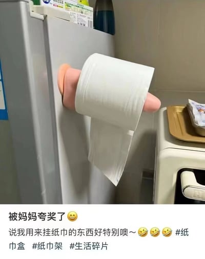 图片展示了一个挂在冰箱侧面的纸巾架：一卷白色纸巾被固定在一个粉色手形支架上，支架吸附在冰箱门上。下方配文描述妈妈夸奖这个挂纸巾的东西“好特别”，带有三个笑哭表情和相关话题标签，整体呈现出日常生活中有趣的小创意场景，充满温馨幽默的氛围。