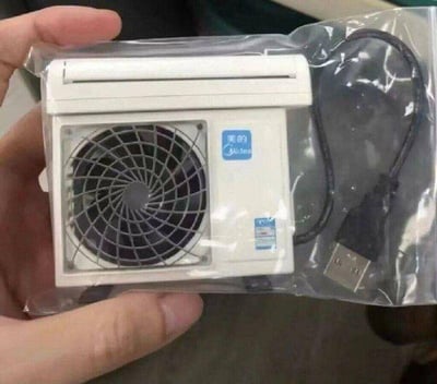这张图片展示了一只手握着被透明塑料包装袋封装的迷你物件，该物件高度还原了美的空调外机的外观细节，包括黑色的风扇格栅、机身右侧的“美的 Midea”品牌标识，还附带一条USB连接线，看起来是一款创意十足的迷你电子小配件，大概率是USB小风扇类产品，整体造型小巧精致，充满趣味，很适合作为桌面小摆件或实用小周边。