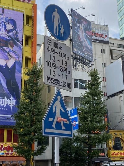 这张图片拍摄于日本繁华的商业街区（从背景的《原神》广告牌推测为东京秋叶原），画面中心是一组立在街头的交通标识：最上方是蓝底白色图案的行人携带儿童通行的指示标志；中间是一块白底黑字的日文告示牌，内容为“周日 4月~9月为13-18点，10月~3月为13-17点（特定禁止区间）”，提示该时段此区域有通行限制；最下方的蓝底白色行人过街预告标志被人恶意恶搞，一个类似手指的物体被贴在行人图案的下半身位置，形成低俗的视觉玩笑，制造出不雅的视觉错觉。背景中可以看到充满商业气息的建筑，左侧建筑墙面上有游戏《原神》中雷电将军的大型宣传广告牌，还有其他各类动漫、商业店铺的招牌，展现出该商圈的热闹氛围。