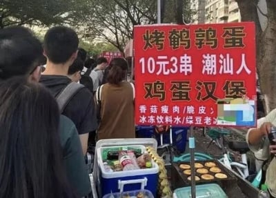 图片展示了一个热闹的街头小吃摊场景。一个红色的招牌十分醒目，上面用白色字体写着“烤鹌鹑蛋 10元3串 潮汕人 鸡蛋汉堡”等字样，还标明了有香菇瘦肉/脆皮、冰冻饮料水/绿豆老冰等小吃饮品。摊位前有不少人在排队购买，能看到各种年龄段的顾客，背景中有树木和建筑物，营造出典型的城市街头美食氛围。