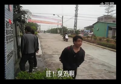 这是一张乡村街道的视频截图，画面中右侧的男子回头朝向镜头方向，表情夸张地张大嘴巴，配合字幕喊出“哇！臭鸡啊”，充满生活化的搞笑感。背景是乡镇的开阔道路，路上有骑电动车的路人，路边分布着民居、厂房，还有行人在墙边停留，整体场景十分接地气。
没有双关、谐音梗或暗示内容，是直白的口语化吐槽场景。