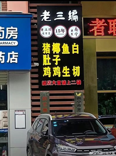 这是一张街头实拍的店铺招牌图，画面中间是一家名为“老三编”的饭店竖版招牌，招牌为黑底，上方标注了“15年”“南宁老友”“老三编家常菜”的字样，中间的菜品名称因排版问题，将“猪肚、椰子鸡、鱼生、白切鸡”拆成了“猪椰鱼白、肚子、鸡鸡生切”的排列方式，形成了容易让人误解的奇怪表述，下方红字提示“酒店大堂请上二楼”。招牌左侧是蓝色的药店店铺，下方停着一辆深色SUV。
招牌因文字排版失误，产生了搞笑的歧义效果，成为核心笑点。