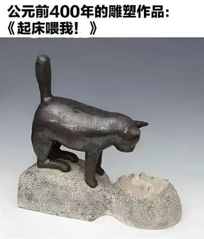 图片展示了一座看起来像公元前400年风格的雕塑作品，雕塑主体是一只猫站立在一个人脸形状的物体上。图片上方配有文字"公元前400年的雕塑作品:《起床喂我!》"，通过将古老雕塑与现代宠物主人常见的经历相结合，创造出幽默效果。这种幽默源于将古代艺术品赋予现代宠物行为的拟人化想象，暗示即使在公元前400年，猫咪可能就已经用这种姿态要求人类喂食了。