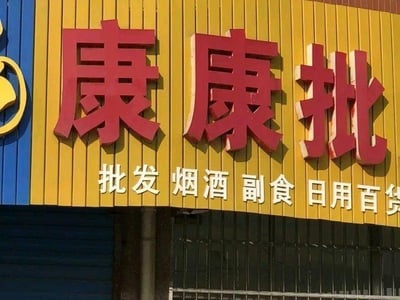 图片展示了一家批发店的招牌。招牌主体为黄色木板拼接背景，上方有红色立体大字“康康批”，下方配有白色小字“批发烟酒副食日用百货”。左侧为蓝色竖条装饰，内含一个黄色圆形图案。整体构图清晰，信息明确，是典型的街边商铺招牌设计。