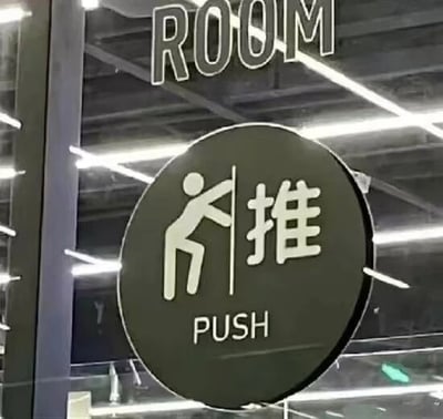 图片展示了一扇玻璃门上的圆形黑色推门标志。标志中央是一个白色简笔画小人，其身体向后大幅度倾斜，双臂伸直向前推，姿势夸张得近乎攀爬，右侧配有白色中文“推”字，下方则是英文“PUSH”字样。背景可见室内天花板的条形灯光和金属结构，整体场景像是商场、办公楼等公共空间的玻璃门区域。

特别之处在于标志中的小人图标姿势过于用力，与常见简洁的推门图标形成反差，显得滑稽有趣，让人联想到“这门是有多难推”的幽默效果。
