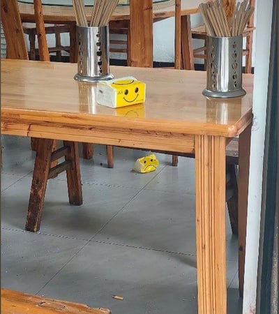 这张图片拍摄于一家餐馆，画面中桌子上和地板上都能看到黄色的纸巾盒。桌上的纸巾盒上有一个可爱的笑脸图案，而地上的纸巾盒则被丢弃，看起来皱巴巴的，但依旧露出了同样的笑脸。

画面形成鲜明对比，桌上纸巾盒“精神饱满”，而被丢弃的纸巾盒虽然境遇不同，却依旧“保持微笑”，带有明显的讽刺和积极的自嘲意味。

**双关/暗示点**：
- 桌下被丢弃的笑脸纸巾盒形成“即使身处低谷也要保持微笑”的暗示，常被网友用来调侃工作、生活等压力话题，象征“表面笑嘻嘻，实际很无奈”。